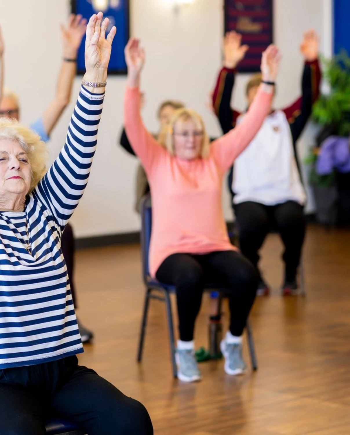 A friendly, inclusive chair-based exercise class — older adults raising their arms together in a bright community hall