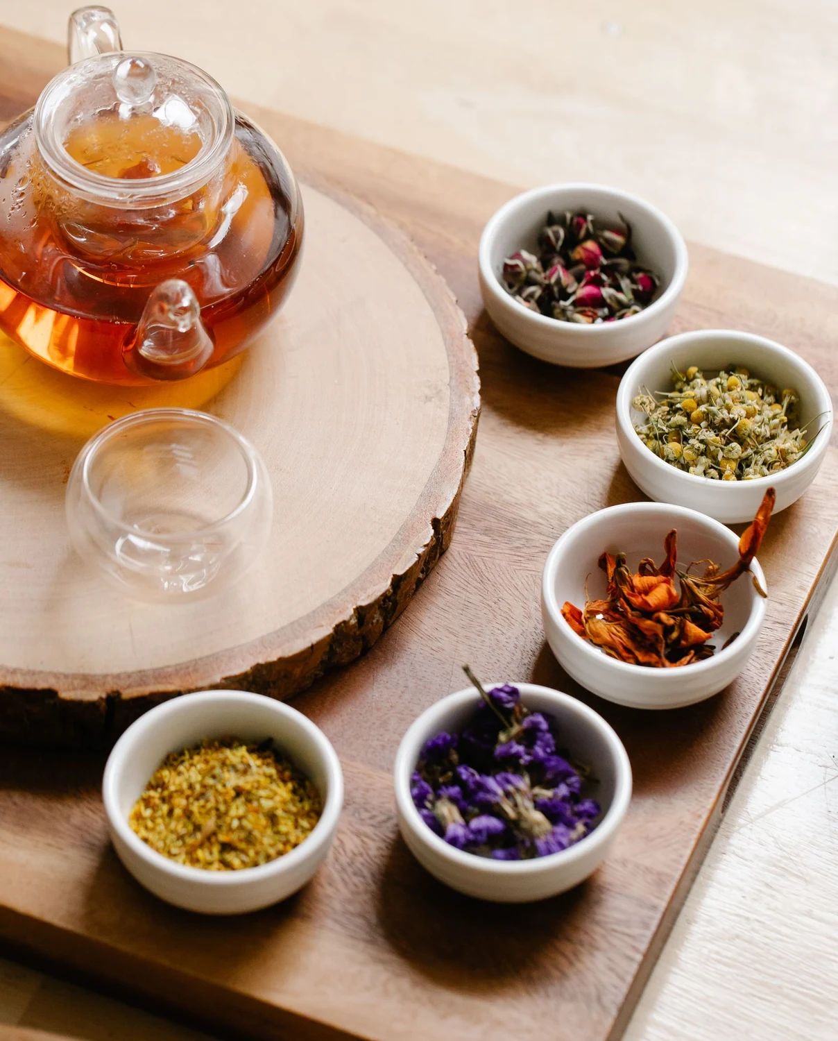 Illustrative atmosphere — a glass teapot and small bowls of dried herbs and botanicals in soft natural light